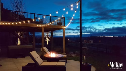Las luces de cuerda de bistro al aire libre crean un ambiente agradable y suave alrededor de un pozo de fuego al aire libre | McKay Landscape Lighting - Omaha, Nebraska